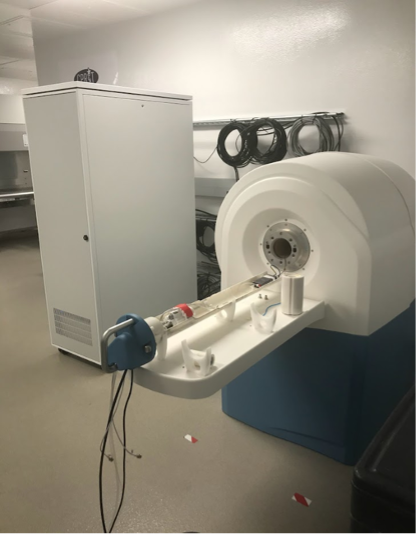 An MRS 3017 MRI machine situated in a clean, organized room. The machine features a cylindrical opening with an attached platform for scanning. In the background, a tall white cabinet and several coiled cables are mounted on the wall.